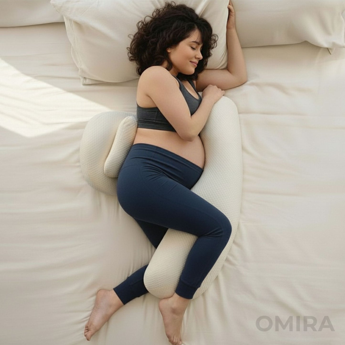 Pregnant woman sleeping on her side with a slim white pregnancy pillow, offering belly and leg support on a bed.