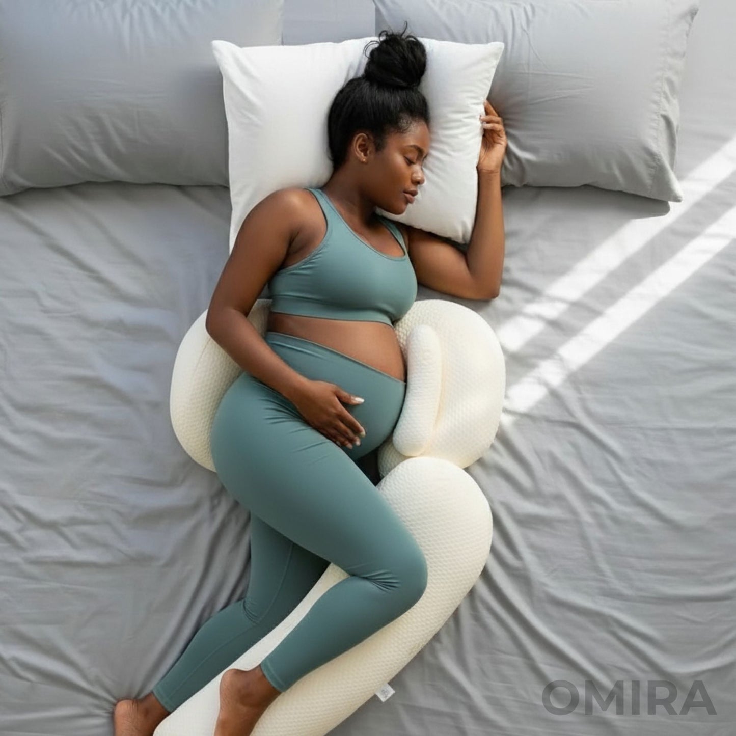 Pregnant woman resting on a grey bed with a white pregnancy pillow, giving belly and leg support for side sleep.